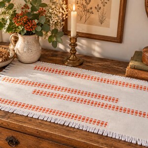 Pode incluir: Um caminho de mesa retangular em tom branco sujo com três filas horizontais de padrões costurados em laranja. O caminho de mesa possui uma borda com franjas nos quatro lados, com as franjas estendendo-se a partir do tecido.