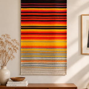 May include: A rectangular woven wall hanging suspended from a wooden rod, featuring a gradient of horizontal stripes in black, vibrant red, orange, yellow, and grey, transitioning from a dark top to a light, neutral-toned bottom edge.