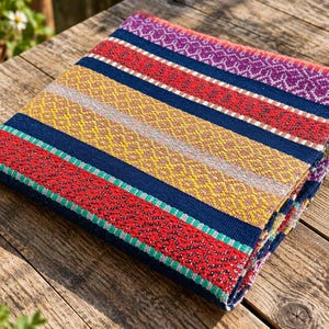 Könnte beinhalten: Ein gefaltetes Textil mit einem lebendigen Muster aus horizontalen Streifen in Marineblau, Rot, Gelb, Lila und Grün. Der Stoff zeigt komplizierte geometrische Webmuster innerhalb jedes farbigen Bandes, was eine strukturierte Optik auf der Oberfläche erzeugt.