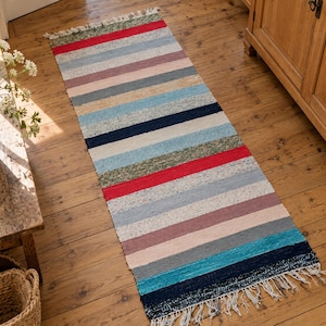 Puede incluir: Una alfombra de pasillo tejida rectangular con un patrón repetitivo de rayas horizontales en varios colores, incluyendo rojo, azul marino, azul claro, gris, rosa y verde. La alfombra tiene una textura tejida y flecos blancos en ambos extremos.