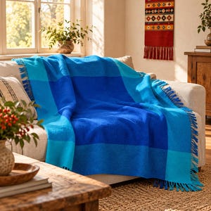 May include: A rectangular throw blanket featuring a large-scale checkered pattern in varying shades of blue, including royal blue, sky blue, and turquoise. The blanket has a textured woven appearance and is finished with fringe along the edges.