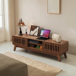 May include: A long, rectangular wooden media console with rounded edges and slatted sliding doors. The top surface holds a brown glass lamp, a framed abstract print, a tablet on a stand, and a small white quilted handbag with a chain strap.