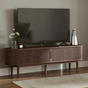 May include: A dark brown wooden media console with a fluted, rounded-edge design and four tapered legs. A flat-screen television sits on top, accompanied by a clear glass vase with white flowers, a small candle, and a silver conical speaker.