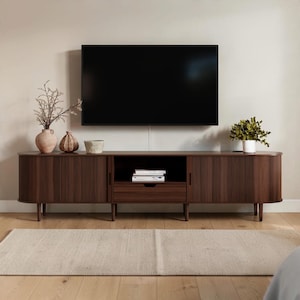 May include: A long, dark brown wooden media console with rounded ends and vertical slatted cabinet doors. It features a central open shelf with a single drawer below, supported by four tapered legs. A flat-screen television is mounted above.