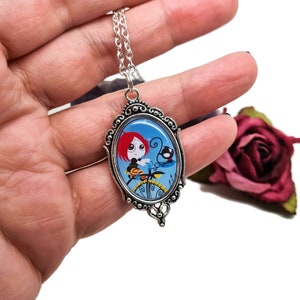 May include: A silver necklace with a blue and white cameo featuring a cartoon girl with red hair and a black bird. The cameo is in an oval frame with ornate details.