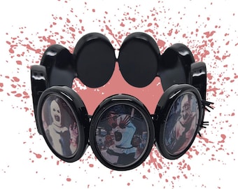 May include: Black bracelet featuring circular images of horror movie characters. The bracelet has a series of black, oval-shaped links. The background has a pink splatter design.