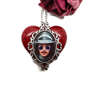 Puede incluir: Un collar con un colgante en forma de corazón rojo con un marco de filigrana plateado. Dentro del marco hay un retrato de un hombre con sombrero y gafas de sol.