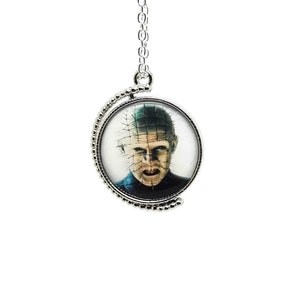Puede incluir: Collar con colgante plateado con un dije circular que muestra el retrato de un personaje de película de terror. El dije tiene un borde con cuentas y una cadena. La imagen muestra un primer plano de la cara del personaje con alfileres.