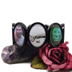 Puede incluir: Pulsera negra con tres dijes ovalados que muestran imágenes de la franquicia de películas de terror Saw. Los dijes muestran a un hombre con la cara ensangrentada, una mano ensangrentada y el muñeco asesino Jigsaw.