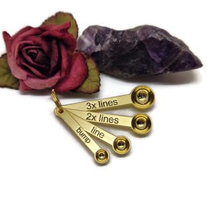 Könnte beinhalten: Satz mit vier goldenen Messlöffeln mit gravierten Markierungen: Beule, Linie, 2x Linien und 3x Linien. Die Löffel sind fächerförmig angeordnet.