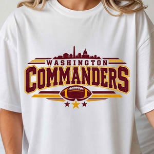 May include: A white t-shirt featuring a graphic of the Washington Commanders logo in burgundy and gold. The design includes a city skyline silhouette, the text WASHINGTON COMMANDERS, a football illustration, and three stars at the bottom.