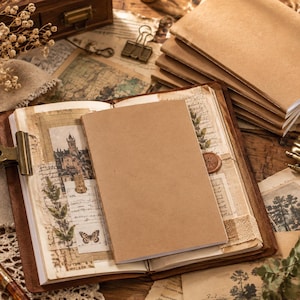 May include: A stack of tan kraft paper notebooks with a central open journal featuring vintage-style collage elements, including botanical illustrations, a butterfly, a building sketch, and handwritten script on aged paper pages.