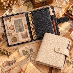 May include: A black crocodile-textured leather binder with a gold six-ring mechanism and a pen, alongside a closed cream-coloured crocodile-textured leather binder with a snap closure, both featuring internal pockets and decorative paper inserts.