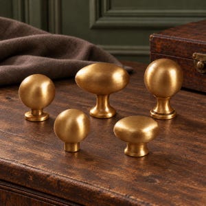 May include: A collection of five gold-toned metal cabinet knobs in various shapes, including spherical, oval, and flattened circular designs, all featuring a smooth, brushed metallic finish and a sturdy base for mounting.