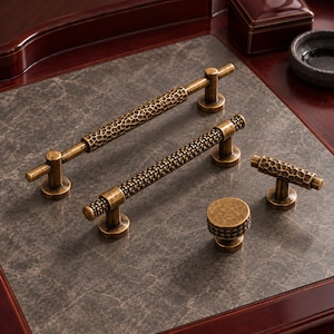 May include: A collection of four antique brass cabinet hardware pieces including two long bar pulls with hammered textures, one smaller T-bar pull with a textured surface, and one round knob with a knurled edge and a weathered finish.