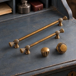 May include: Two long, slender cabinet pulls and two round cabinet knobs made of antique brass-toned metal. The pulls feature fluted, fan-shaped ends and a smooth central bar, while the knobs have a matching fluted or concentric ring design.