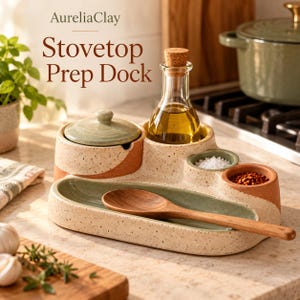 Op de afbeelding: Een keramische Stovetop Prep Dock van AureliaClay met een gespikkelde crème, saliegroene en terracotta afwerking. Het bevat een bakje met deksel, een glazen oliekaraf met kurk, twee kleine bakjes voor kruiden en een houten lepel.