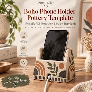 May include: A beige speckled ceramic phone holder with a flat base and angled front slot. The surface features painted terracotta, dark green, and black motifs including arches, leaves, and a circle. A white charging cable is inserted at the bottom.