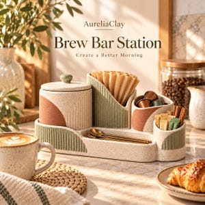 May include: A ceramic coffee station organizer featuring a speckled cream finish with sage green and terracotta accents. The set includes a lidded canister, a filter holder, a compartment for coffee pods, and a tray for gold spoons.