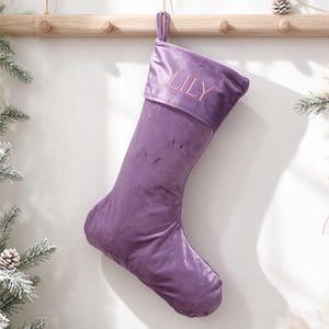 May include: A purple velvet Christmas stocking featuring a folded cuff at the top. The name LILY is embroidered in light pink thread on the centre of the cuff. A matching purple fabric loop is attached to the top for hanging purposes.