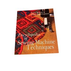 May include: A book titled Encyclopedia of Sewing Machine Techniques by Nancy Bednar and JoAnn Pugh-Gannon. The cover features a close-up of a sewing machine presser foot stitching a red, blue, and black patterned fabric onto gold fabric.