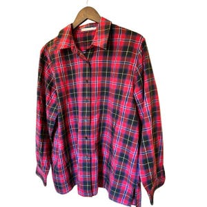May include: A long-sleeve button-down shirt featuring a vibrant red, black, and white plaid pattern. The shirt has a classic collar, a full-length front button placket, and long sleeves with buttoned cuffs, all displayed on a wooden hanger.
