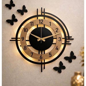 May include: A round wall clock featuring a black center, a light wood-toned ring with black numerals from 1 to 12, and gold-colored hands. The clock is framed by black geometric lines and illuminated by a warm backlight, accompanied by five black butterfly wall decals.