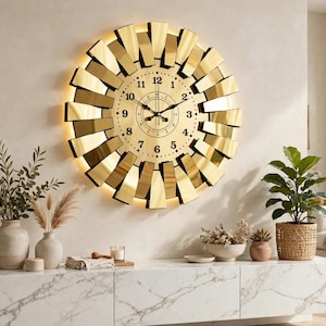 May include: A large circular wall clock with a gold-toned face and black numerals. The clock features a sunburst-style frame made of reflective gold-colored segments arranged in a radial pattern, with integrated backlighting behind the frame.