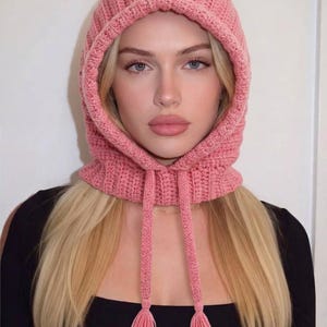 May include: A dusty pink crochet balaclava hood featuring a ribbed knit texture, a rounded head shape, and two long hanging ties at the front, each finished with a small matching tassel at the end. The hood is constructed from yarn.