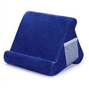 May include: A royal blue triangular tablet stand made of soft, plush fabric. The stand features a wide base with a built-in ledge to support a device and a small white mesh pocket on the side for storage. The design is angled for viewing.