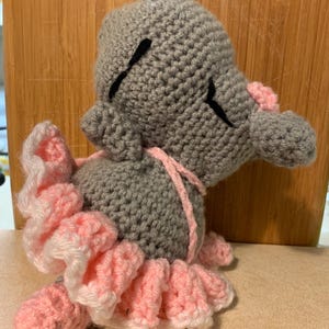 May include: A gray crocheted stuffed animal featuring a pink ruffled skirt and a small pink nose. The figure has closed eyes represented by black stitched lines and is constructed from yarn with a textured, looped surface throughout the body.