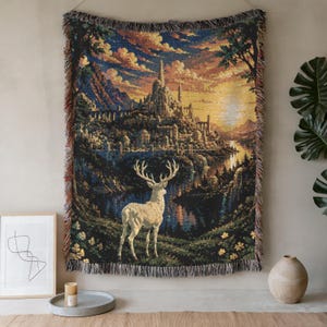 May include: A woven tapestry blanket featuring a detailed scene of a white stag standing in a grassy meadow, overlooking a river and a large castle complex nestled among mountains under a golden sunset sky, finished with a fringed edge.