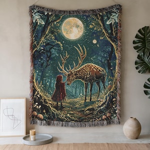 May include: A woven tapestry blanket featuring a dark forest scene at night with a large full moon, a person in a red hooded cloak, and a large stag with antlers. The rectangular textile has fringed edges and a detailed, colorful thread design.