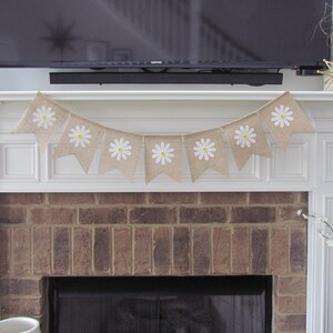 May include: A burlap banner with white daisy flowers and yellow centers hangs above a brick fireplace. The banner is strung with white twine and has a scalloped edge.