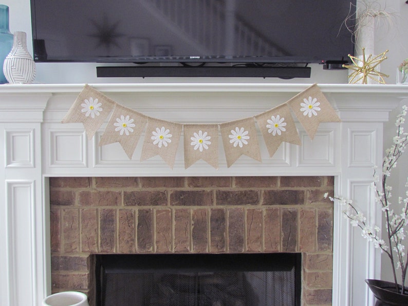 May include: A burlap banner with white daisy flowers and yellow centers hangs above a brick fireplace. The banner is strung with white twine and has a scalloped edge.