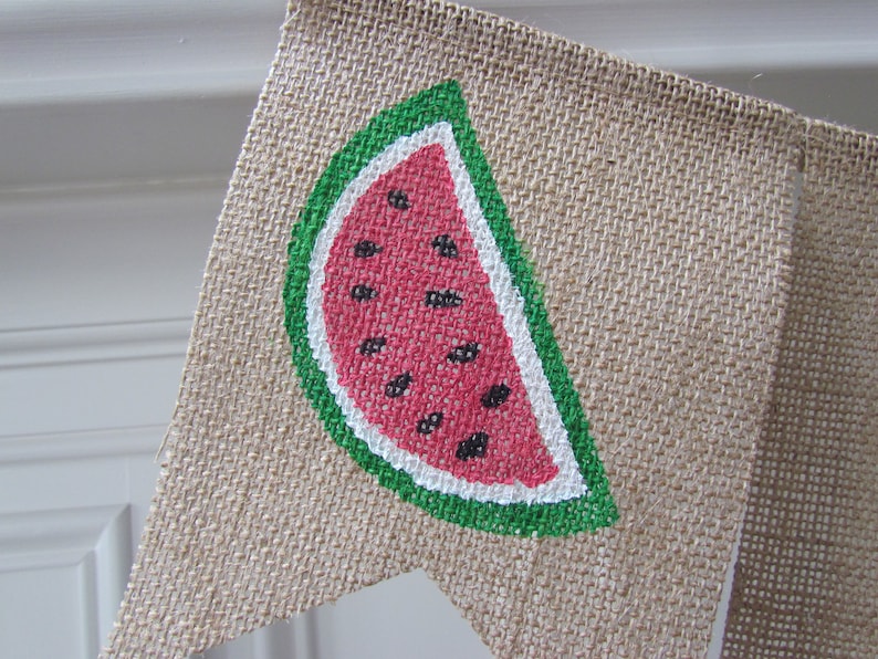 May include: A burlap banner with a painted watermelon slice. The watermelon is red with black seeds and a green rind.