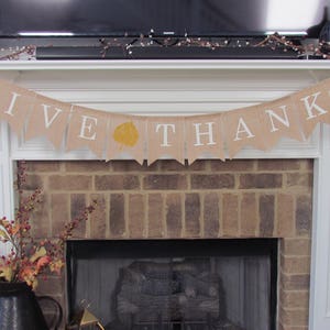 May include: A burlap banner with the words "GIVE THANKS" in white lettering hangs above a brick fireplace. A single yellow leaf is printed on the banner.