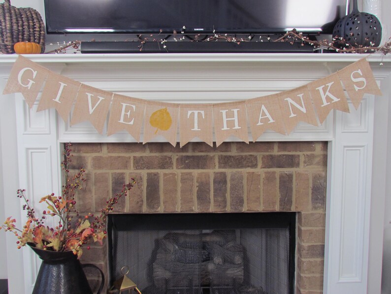 May include: A burlap banner with the words "GIVE THANKS" in white lettering hangs above a brick fireplace. A single yellow leaf is printed on the banner.