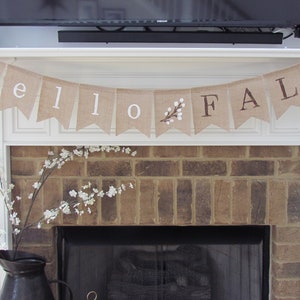May include: A burlap banner with the words "hello fall" hanging above a brick fireplace. The banner has white lettering and a small sprig of white flowers in the center.
