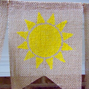 May include: A burlap banner with a yellow sun painted on it. The banner is a triangle shape and has a frayed edge.