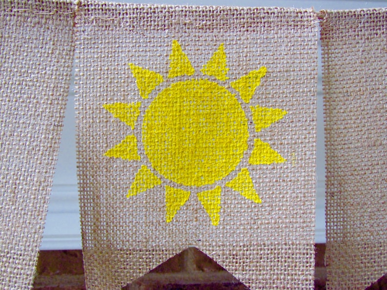 May include: A burlap banner with a yellow sun painted on it. The banner is a triangle shape and has a frayed edge.