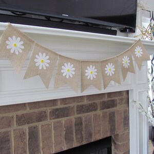 May include: A burlap banner with white daisies and yellow centers hangs above a brick fireplace. The banner is strung with white twine and has a rustic, farmhouse style.
