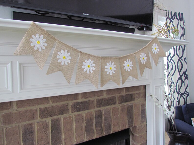 May include: A burlap banner with white daisies and yellow centers hangs above a brick fireplace. The banner is strung with white twine and has a rustic, farmhouse style.