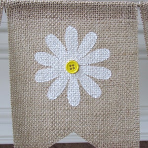May include: A burlap banner with a white painted daisy and a yellow button in the center. The banner is part of a garland.