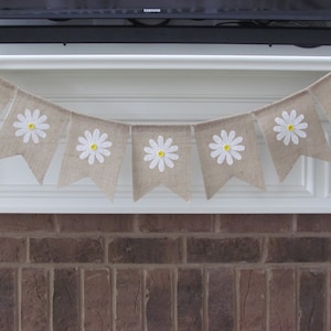 May include: A burlap banner with white daisy flowers and yellow centers. The banner is strung across a white mantel above a brick fireplace.