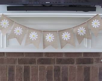 May include: A burlap banner with white daisy flowers and yellow centers. The banner is strung across a white mantel above a brick fireplace.