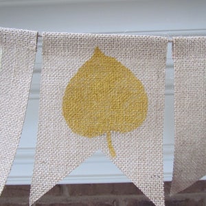 May include: A burlap banner with the word "Fall" in white paint. The letter "A" is decorated with a yellow leaf.