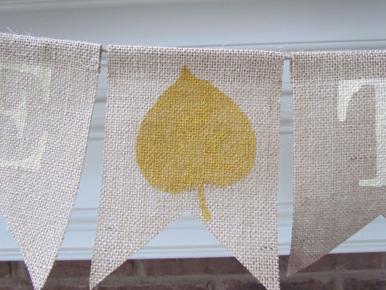 May include: A burlap banner with the word "Fall" in white paint. The letter "A" is decorated with a yellow leaf.