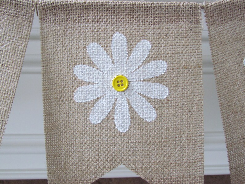 May include: A burlap banner with a white painted daisy and a yellow button in the center. The banner is part of a garland.