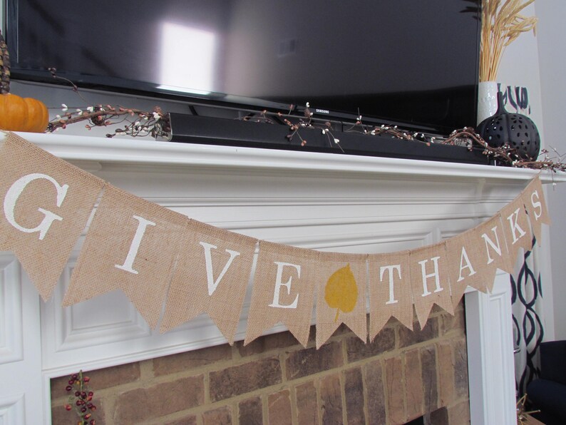 May include: A burlap banner with the words "Give Thanks" in white lettering. The banner is decorated with a yellow leaf and is hanging above a brick fireplace.